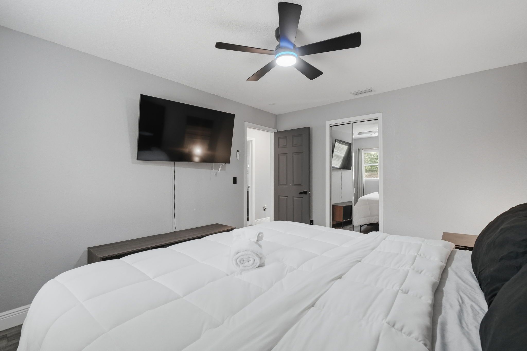 A bedroom with a bed, a ceiling fan, a television mounted on the wall, and a door leading to another room.
