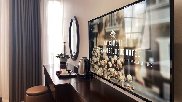A hotel reception area with a large screen displaying the hotel's name and tagline. There is a wooden desk with a kettle, a tray with a teapot, and a potted plant. A mirror hangs on the wall, and a brown ottoman is visible. - Elysinam Boutique Hotel (Bai Chay)