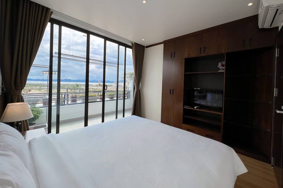 Quarto de hotel com uma cama grande, um guarda-roupa e vista para uma ponte e paisagem urbana através de grandes janelas.