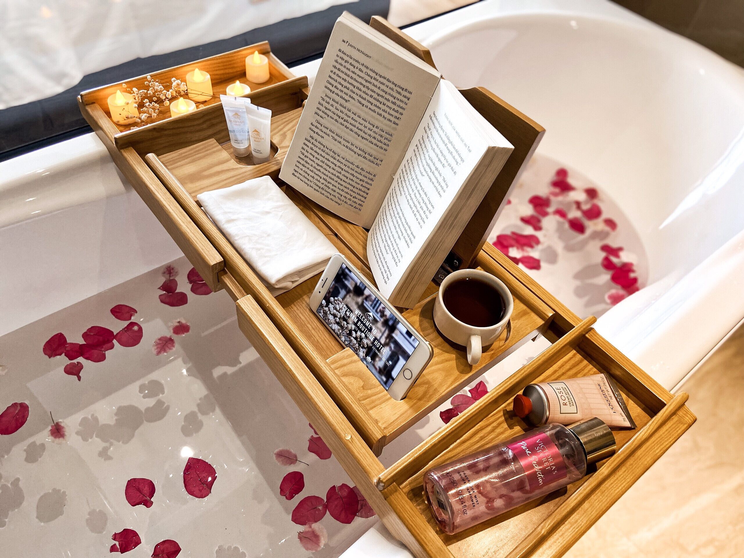 Ein hölzernes Tablett mit Kerzen, einem Buch, einem Smartphone, einer Tasse Tee und Toilettenartikeln auf einer Badewanne voller Rosenblätter.