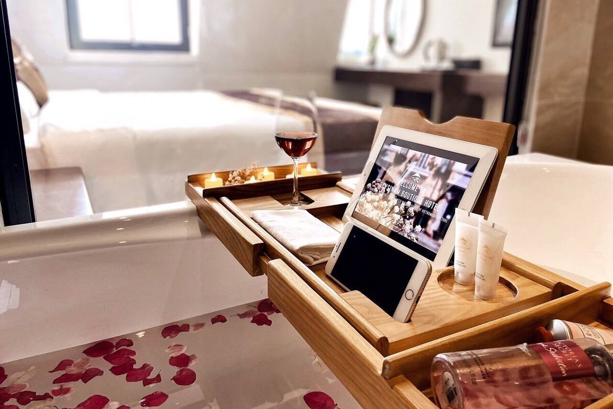 Uma bandeja de madeira com um tablet, um copo de vinho tinto e duas embalagens de loção sobre uma cama com pétalas de rosa.