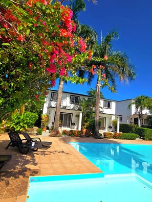 Un espace aménagé au bord d’une piscine à l’eau d’un bleu limpide, avec des palmiers et un bâtiment blanc doté d’un balcon.