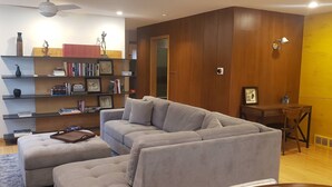 Ein modernes Wohnzimmer mit einem grauen Ecksofa, einem Bücherregal aus Holz und einem kleinen Holztisch mit einem Stuhl.
