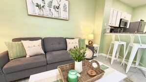Un salon avec un canapé gris, des coussins verts et blancs, une table basse blanche ornée d’une bougie verte et un plateau en osier.