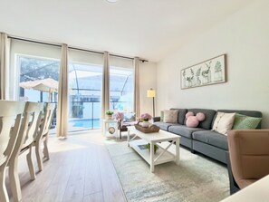 A modern living room with a grey sofa, white coffee table, and a large window overlooking a pool.