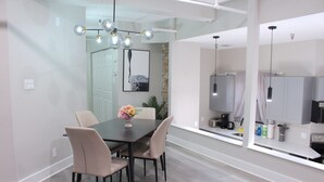 Un moderno comedor con una mesa de madera oscura, sillas color beige y un jarrón de flores. Hay una pared de vidrio que separa el comedor de la cocina, la cual cuenta con gabinetes blancos y un refrigerador de acero inoxidable.