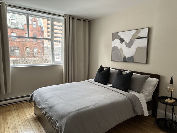 Ein modernes Hotelzimmer mit einem großen Bett, Blick auf die Stadt durch ein Fenster und einer minimalistischen Kunstausstellung an der Wand.