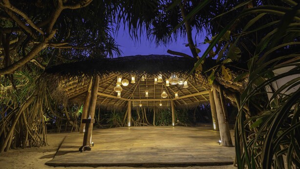 A covered outdoor area with a wooden floor, surrounded by trees and plants, and illuminated by hanging lights. - Jungle Beach Ahungalla - Classic Room (Ahungalla)