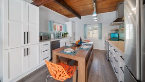 A modern kitchen with white cabinets, a wooden ceiling, and a dining area with a wooden table and orange chairs. - NEW Steps to Beach, Prime City Center, Firepit, Hot Tub, PS5, Pet Friendly (Rockaway Beach)