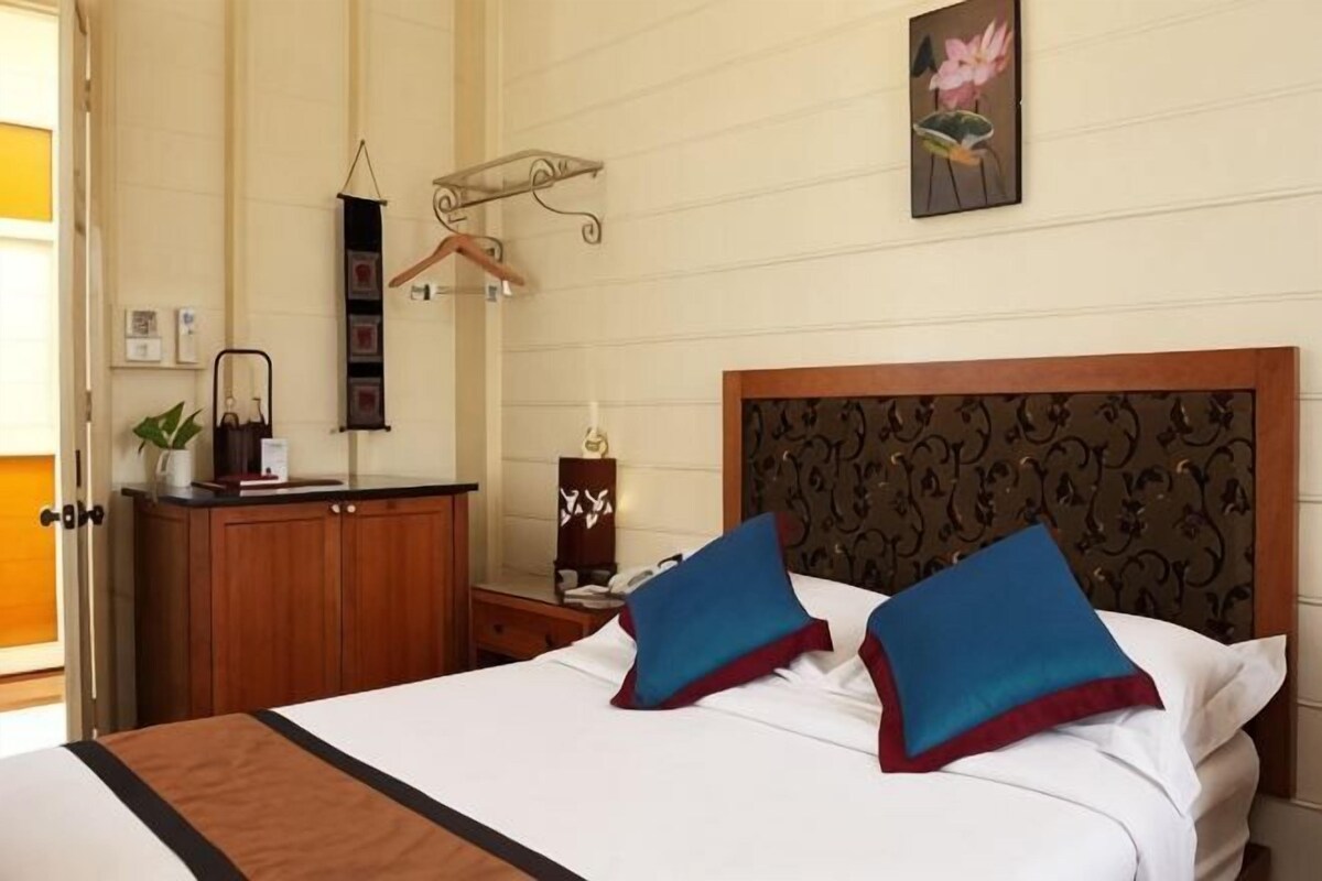 A hotel room with a bed, a wooden headboard, a bedside table, a nightstand, a hanging light fixture, and a wall-mounted clock.