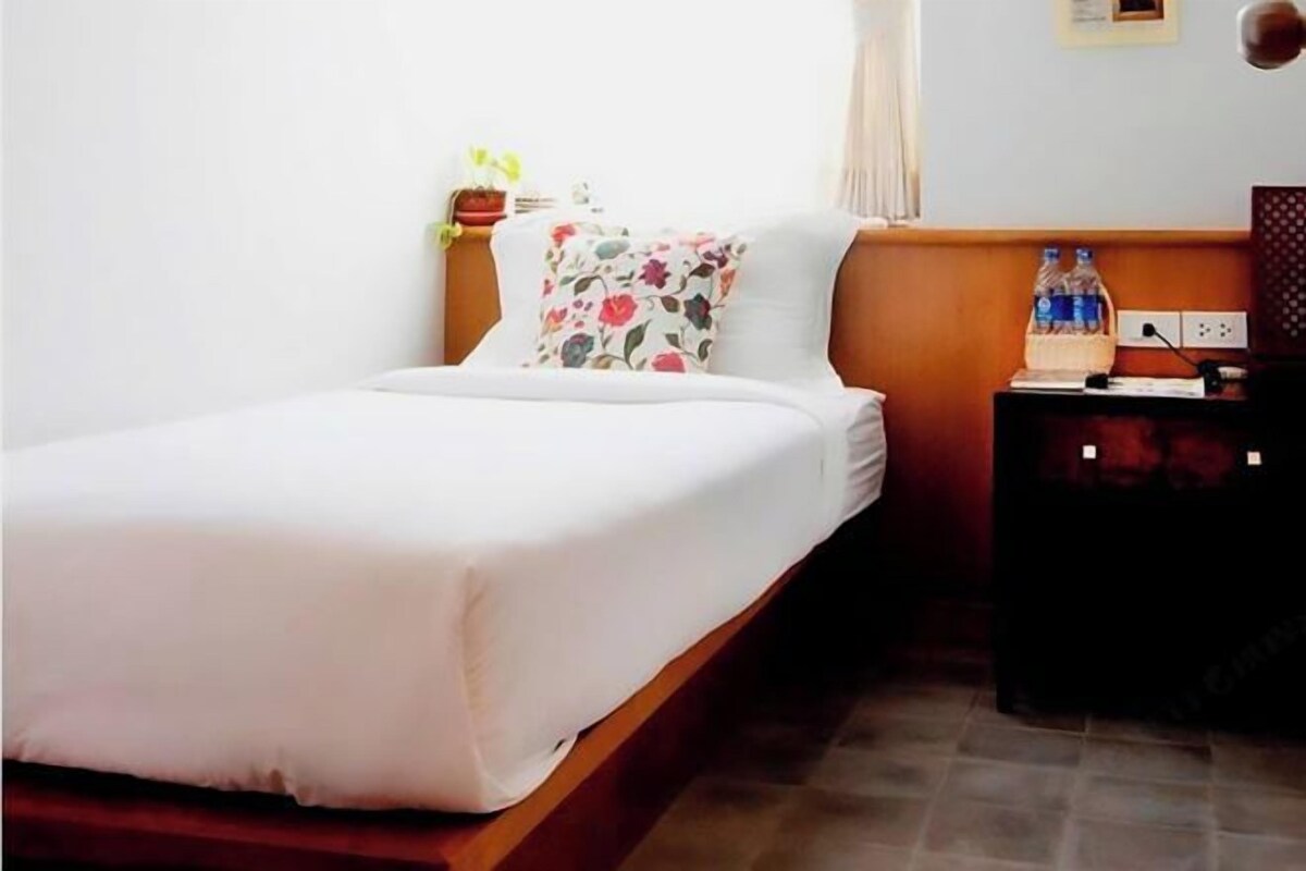A neatly made bed with a floral pillow, a small bedside table with a water bottle, and a power outlet on the wall.