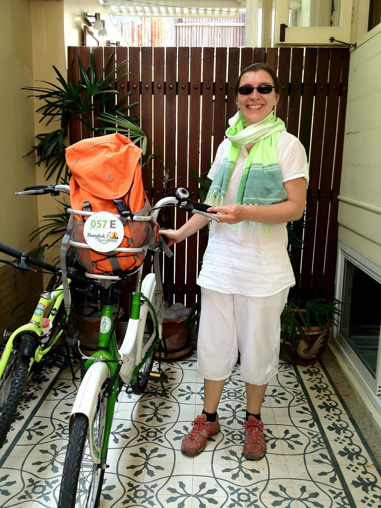 A person standing next to a green bicycle with a basket, in a tiled area with a wooden fence and potted plants.