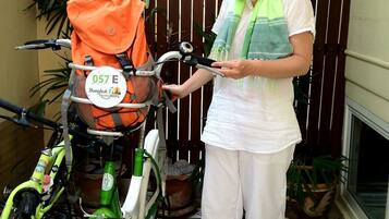 A person standing next to a green bicycle with a basket, in a tiled area with a wooden fence and potted plants.