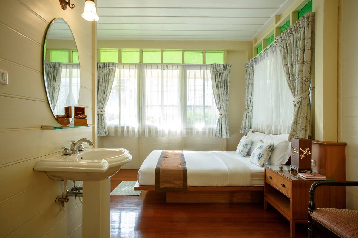 A bedroom with a bed, a wooden nightstand, a mirror, a sink, and a window with curtains.