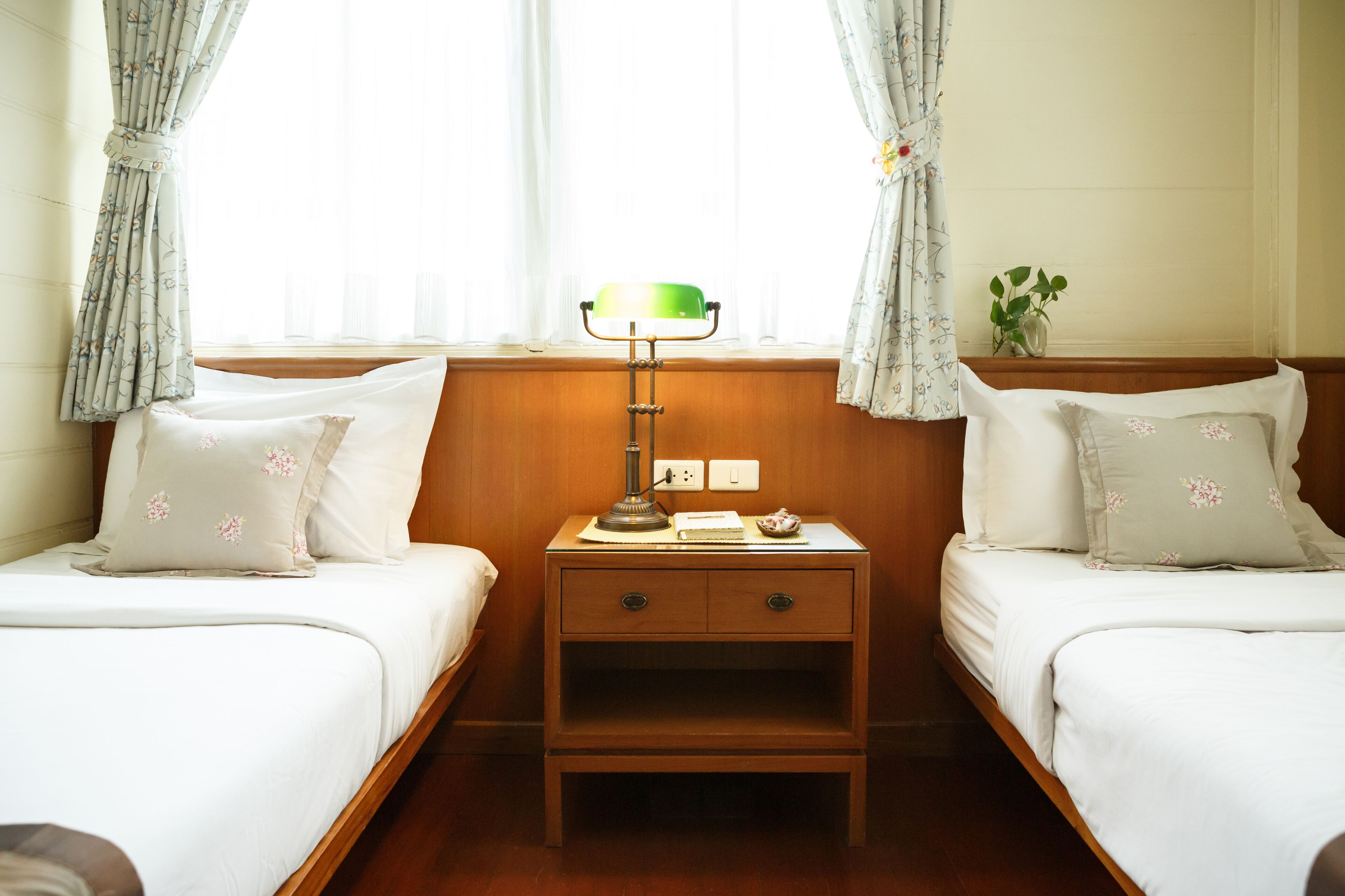 A hotel room with two single beds, a nightstand with a lamp, and a small plant on the nightstand.