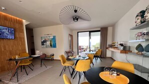 Una habitación de hotel moderna con zona de comedor que presenta sillas amarillas y una mesa negra, un ventanal con vistas y un ventilador de techo.