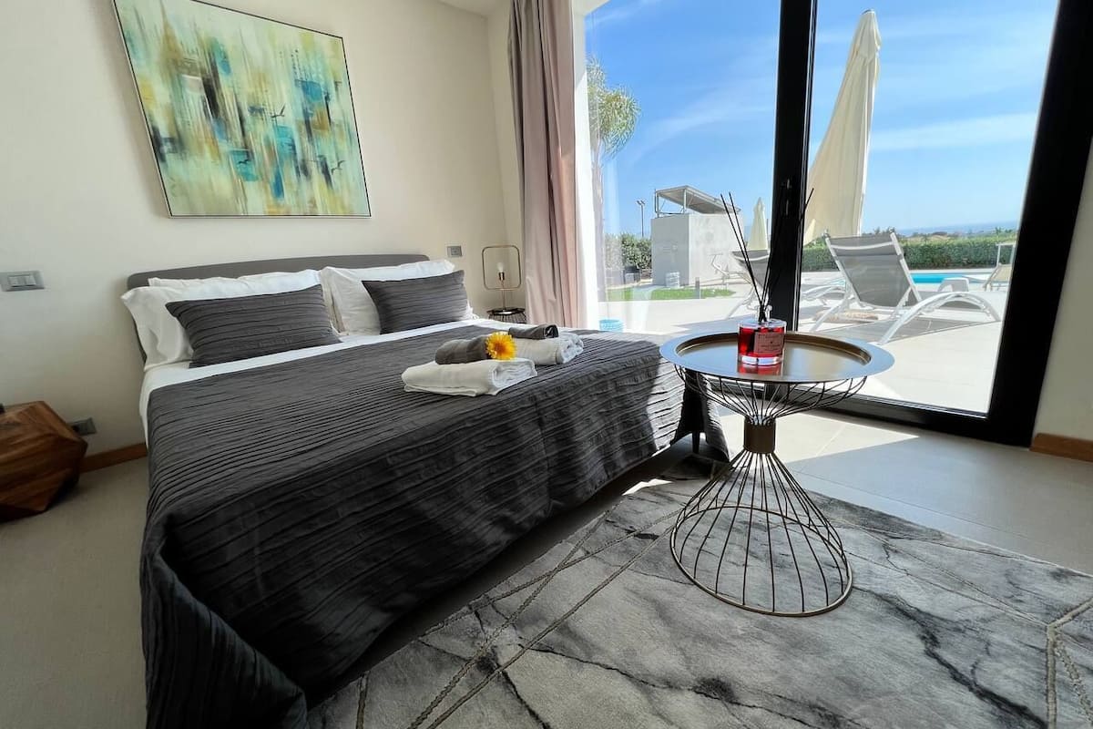 Ein modernes Schlafzimmer mit einem großen Bett, einem runden Couchtisch und Blick auf einen Pool und Außenbereich.