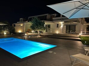 A modern outdoor swimming pool area with a well-lit pool, lounge chairs, and umbrellas.