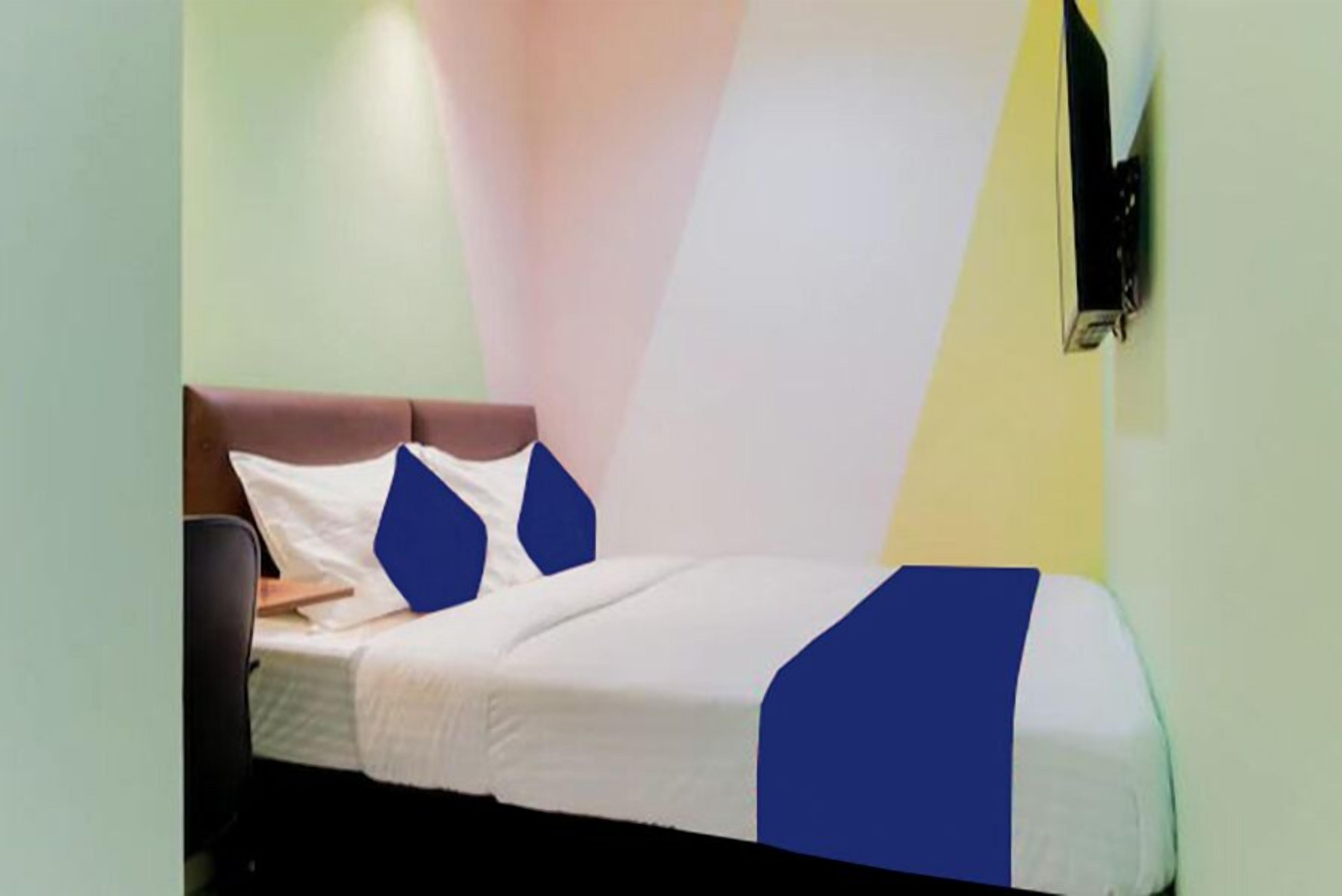 Una cama individual con colcha blanca y azul, cabecera de madera y televisor montado en la pared.