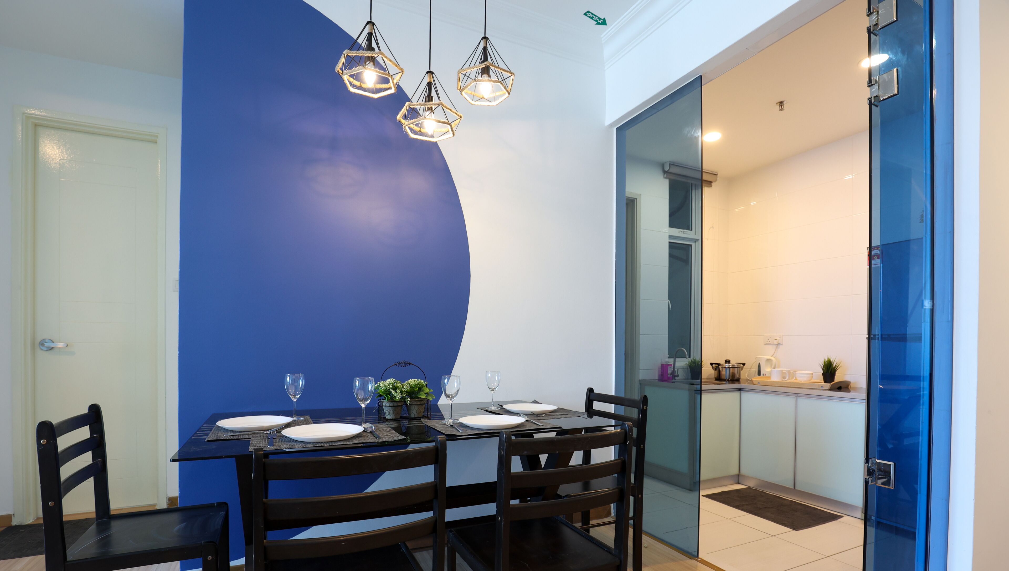 A dining area with a blue accent wall, a glass door leading to a kitchen, and a table set for four with wine glasses and plates.
