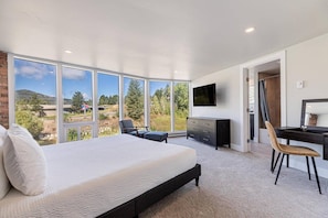 A bedroom with a large bed, a desk, a chair, a TV, and a view of the outdoors.