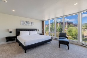 A bedroom with a large bed, a chair, and a view of buildings through large windows.