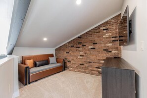 A loft-style room with a sloped ceiling, a brick accent wall, a sofa, and a flat-screen TV mounted on the wall.