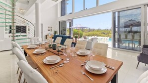 Un'ampia sala da pranzo con un lungo tavolo in legno apparecchiato per un pasto, una moderna scala con ringhiere in vetro e vista su una piscina e vegetazione all'esterno.