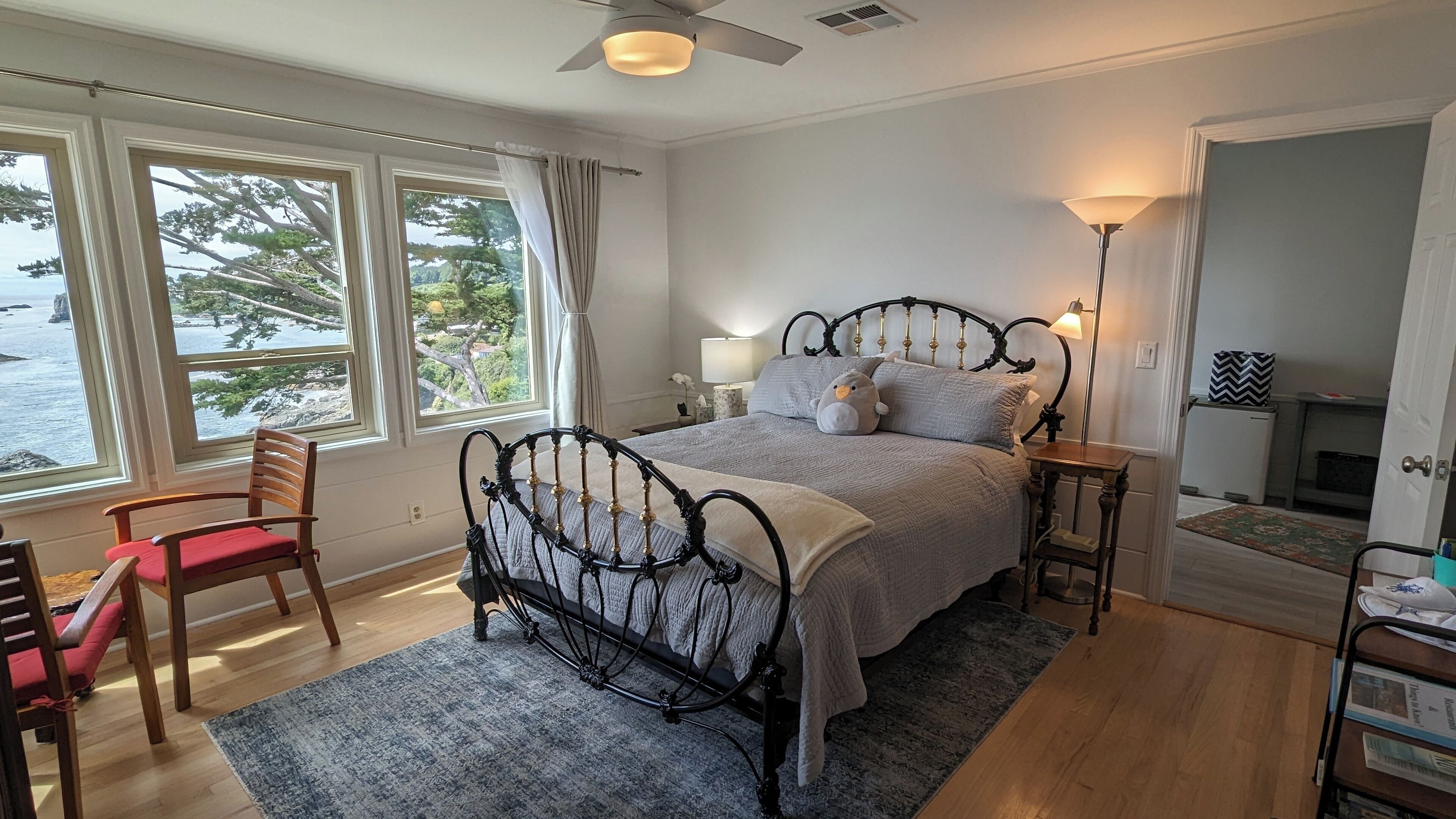 A bedroom with a bed, a chair, a nightstand, a lamp, and a view of the sea through large windows.