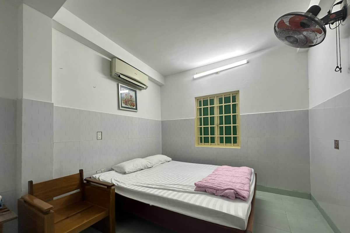 A small, clean room with a single bed, a wooden bench, a window with a green frame, and an air conditioning unit.
