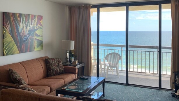 A living room with a sofa, a glass coffee table, and a painting on the wall. There is a view of the ocean through large sliding doors. - Cozy oceanfront beach condo full of light (North Myrtle Beach)
