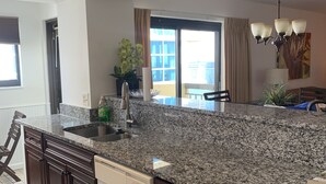 A modern kitchen with granite countertops, dark wooden cabinets, a built-in dishwasher, and a large window offering a view of the outdoors. - Cozy oceanfront beach condo full of light (North Myrtle Beach)
