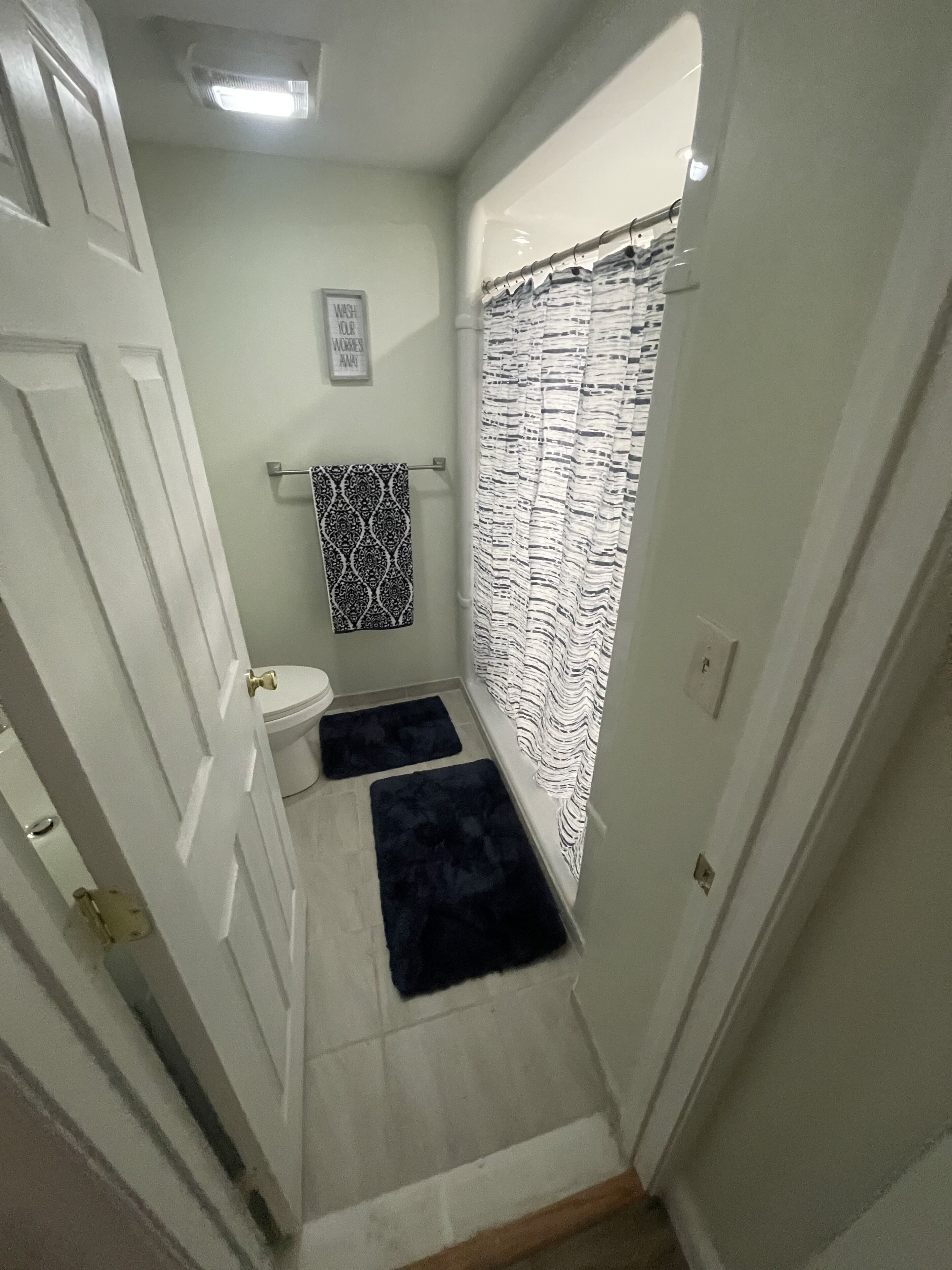 A narrow bathroom with a toilet, a sink, a towel rack with a patterned towel, a shower curtain with a striped pattern, and two dark mats on the floor.