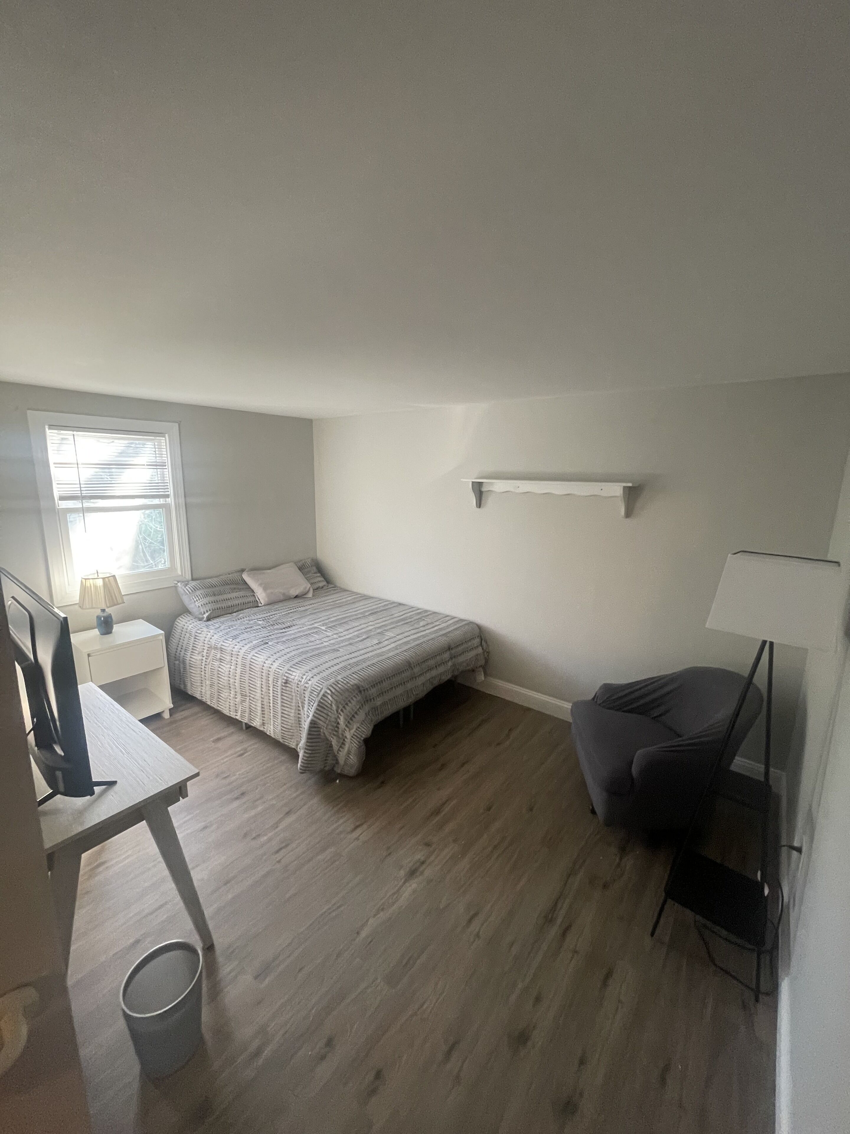 A small, sparsely furnished room with a bed, a desk, a chair, and a window.