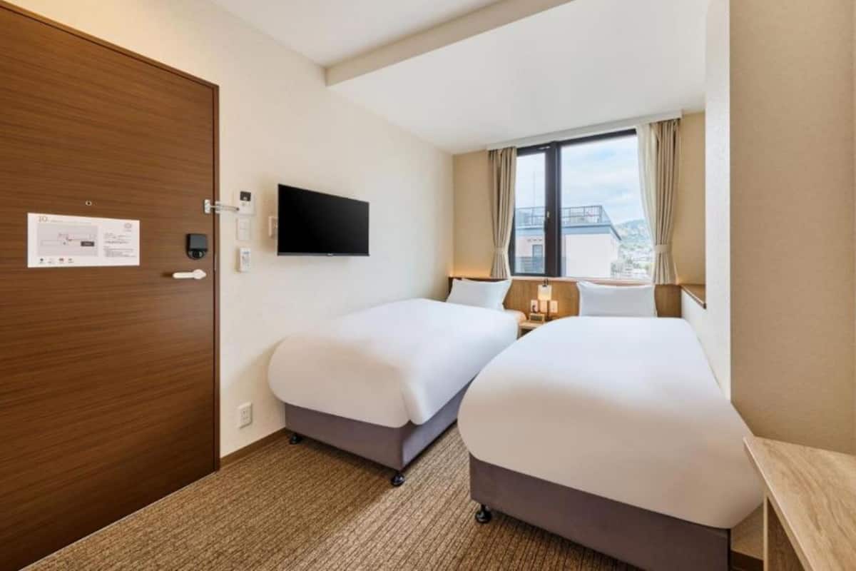 Ein Hotelzimmer mit zwei Betten, einem Flachbildfernseher, einem Fenster mit Vorhängen und Blick auf die Stadt.