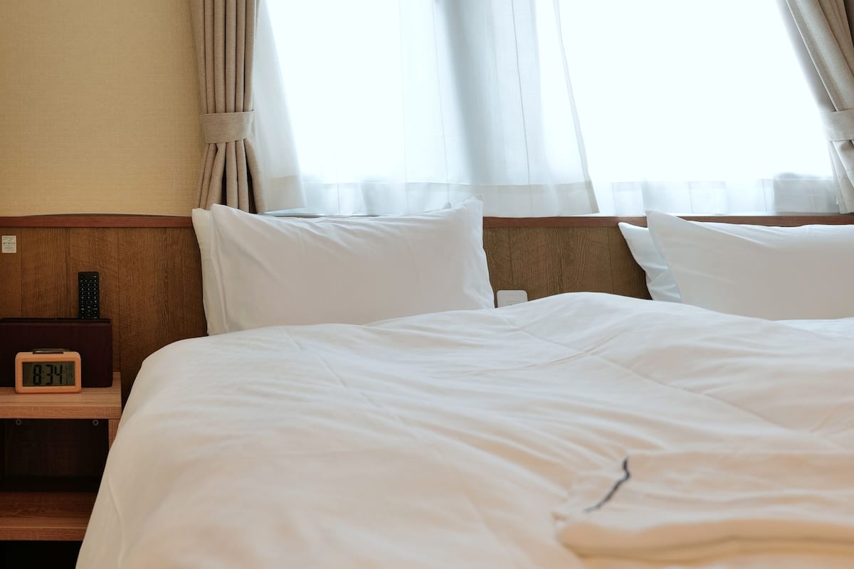 Ein Hotelzimmer mit einem großen Bett, weißen Leinentüchern, einem Holz-Kopfende, einem Nachttisch mit einer Uhr und Fernbedienung sowie transparenten Vorhängen.