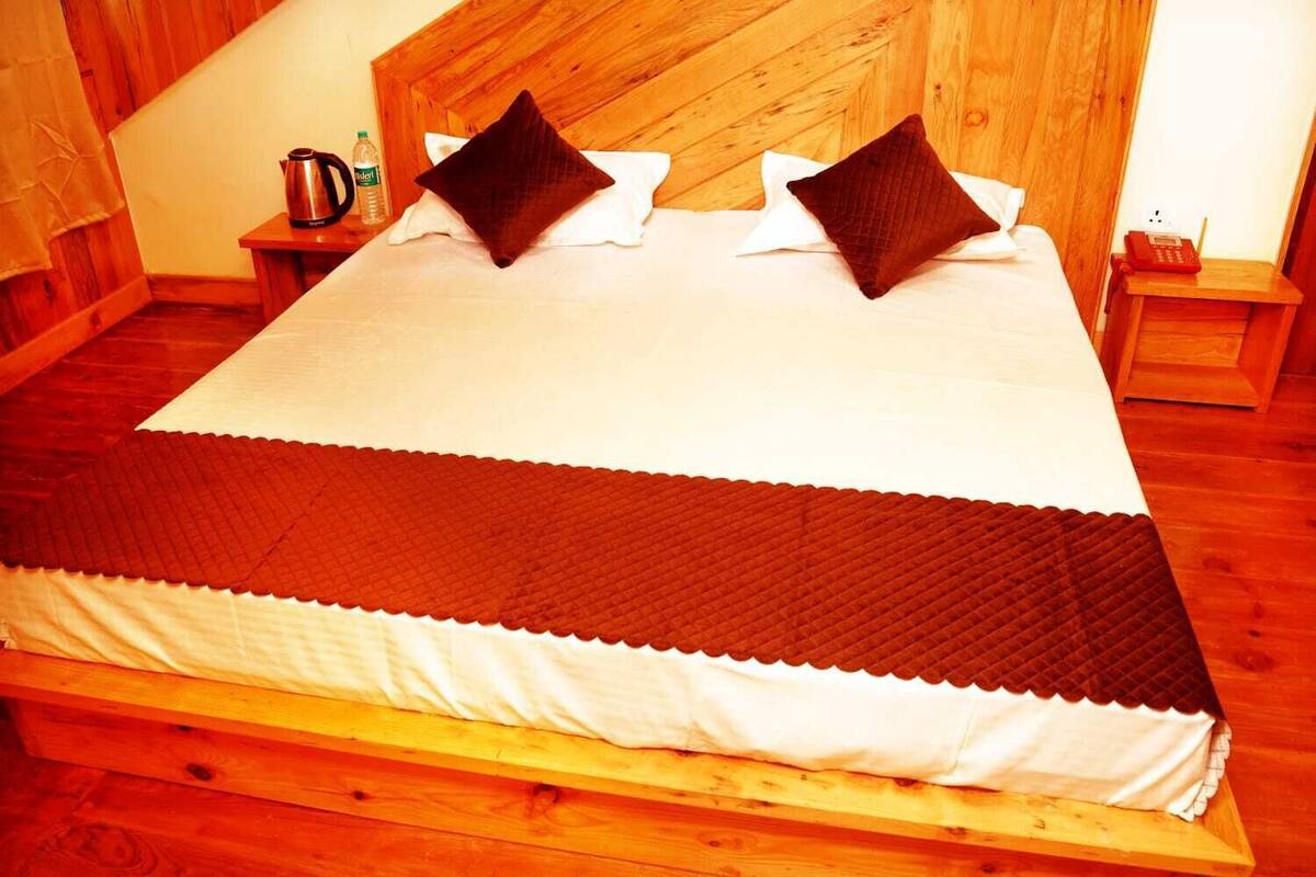 A wooden bed with a white and brown quilt, two pillows, a bedside table with a kettle and a bottle, and a wooden nightstand with a phone.