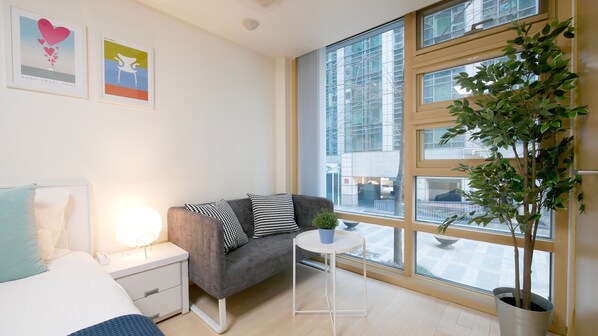 A modern bedroom with a bed, bedside table, lamp, grey sofa with patterned cushions, a small white table with a plant, and a large window with a view of buildings and scaffolding.