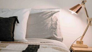 En pænt redt seng med hvidt og gråt sengetøj, en sort pude og et trænatbord med en lampe.