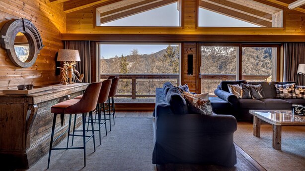A cozy cabin interior with a wooden interior, a large window overlooking a mountainous landscape, a sofa, and a bar area with stools. - Chalet Flocon de Neige - jacuzzi - sauna - La Tzoumaz, 4 Vallees (La Tzoumaz)