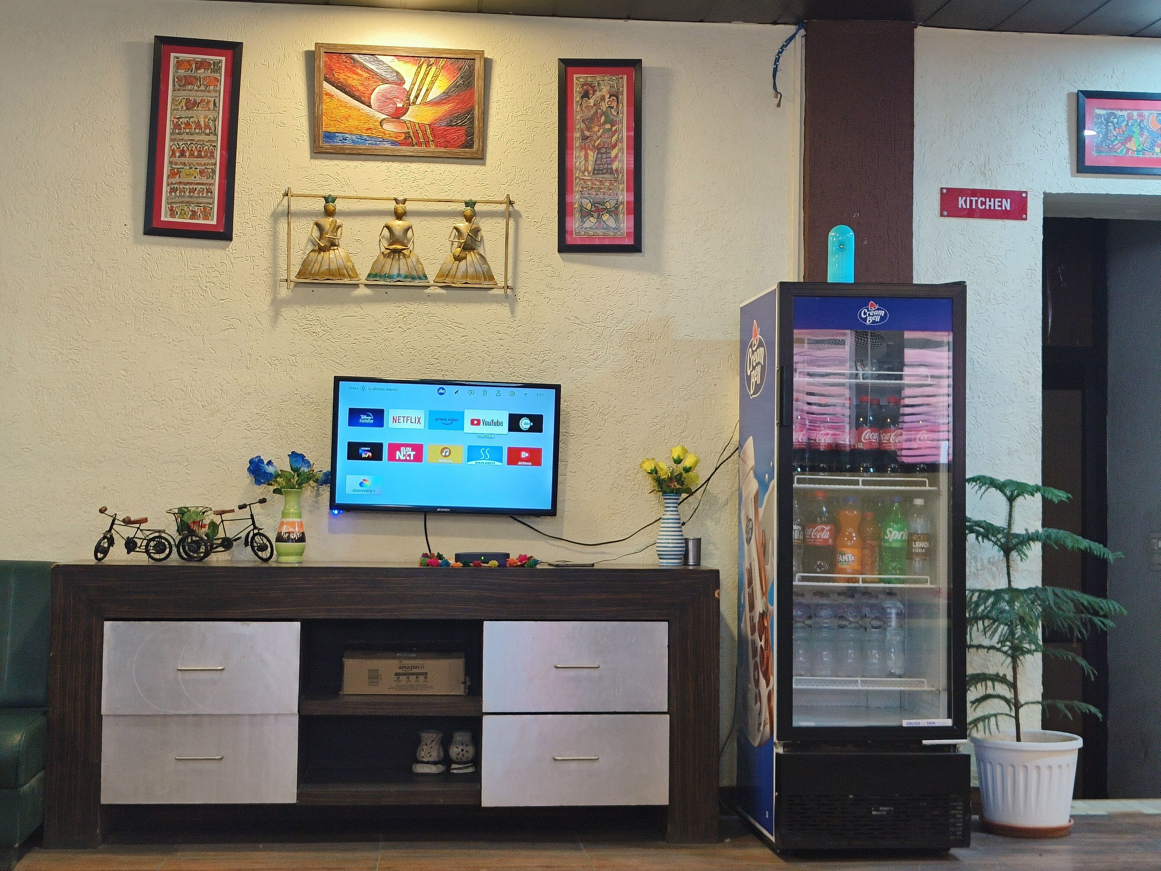 A hotel lobby with a television displaying various streaming services, a refrigerator vending machine, and a reception desk with a vase of flowers.