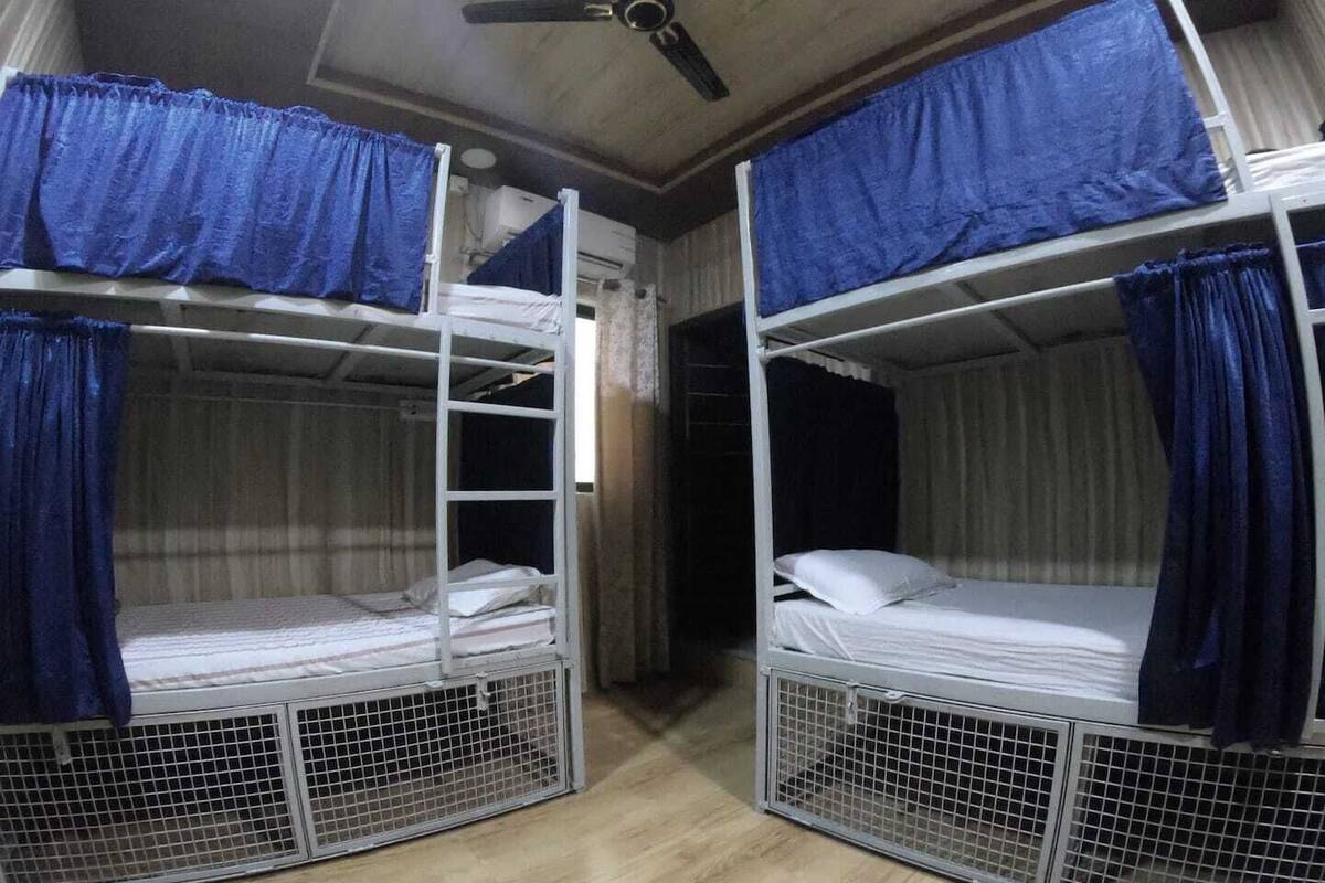 Quarto com duas beliches, cada uma com estrutura de metal e cortinas azuis. Há um ventilador de teto e uma janela com cortinas.
