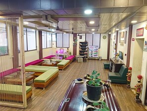 Une pièce avec un plancher en bois, des sièges rembourrés de couleur verte et rose, une table basse en bois, une bibliothèque, une petite table avec une plante et un téléviseur posé sur un bureau.