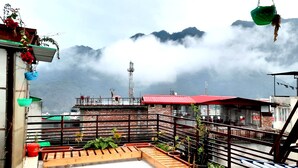 Una terraza en la azotea con plantas en macetas, un edificio de techo rojo y un paisaje montañoso de fondo.