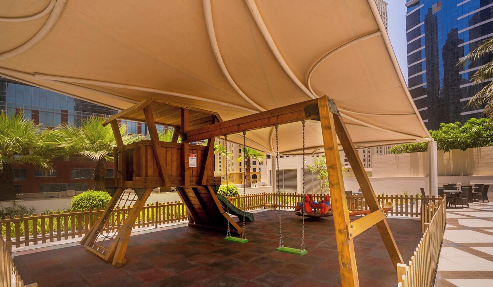 Un ensemble de balançoires en bois, placé sous une sorte d’auvent et entouré d’une clôture, avec des bâtiments en arrière-plan.
