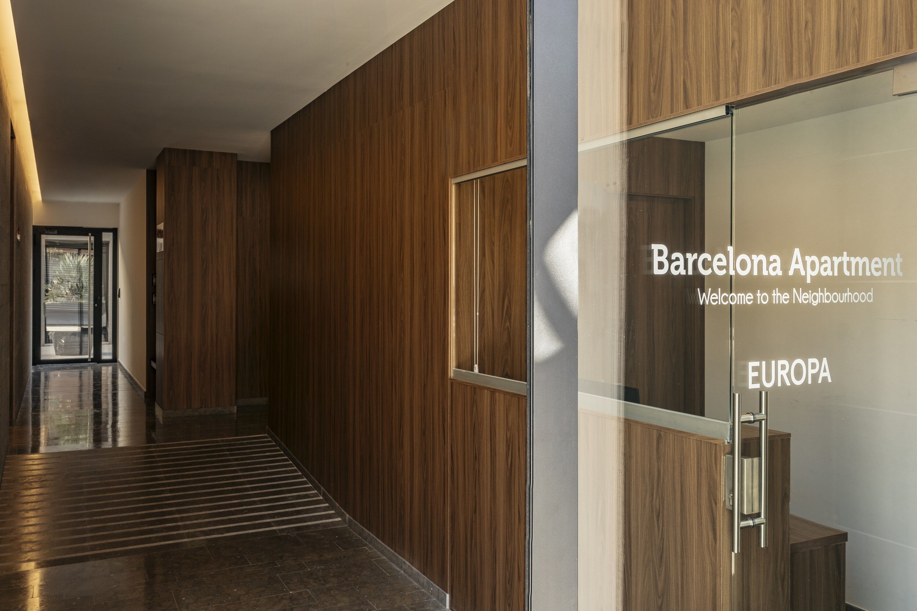 A modern hallway with wooden paneling and a glass door with text indicating a Barcelona apartment.
