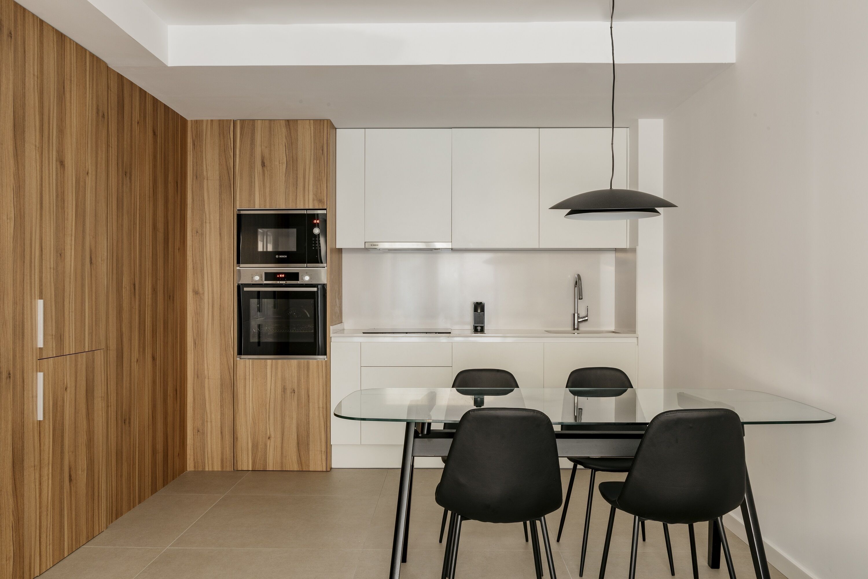 Una cocina moderna con un armario de madera, un horno Bosch, una encimera blanca y una mesa de comedor de vidrio con sillas negras.