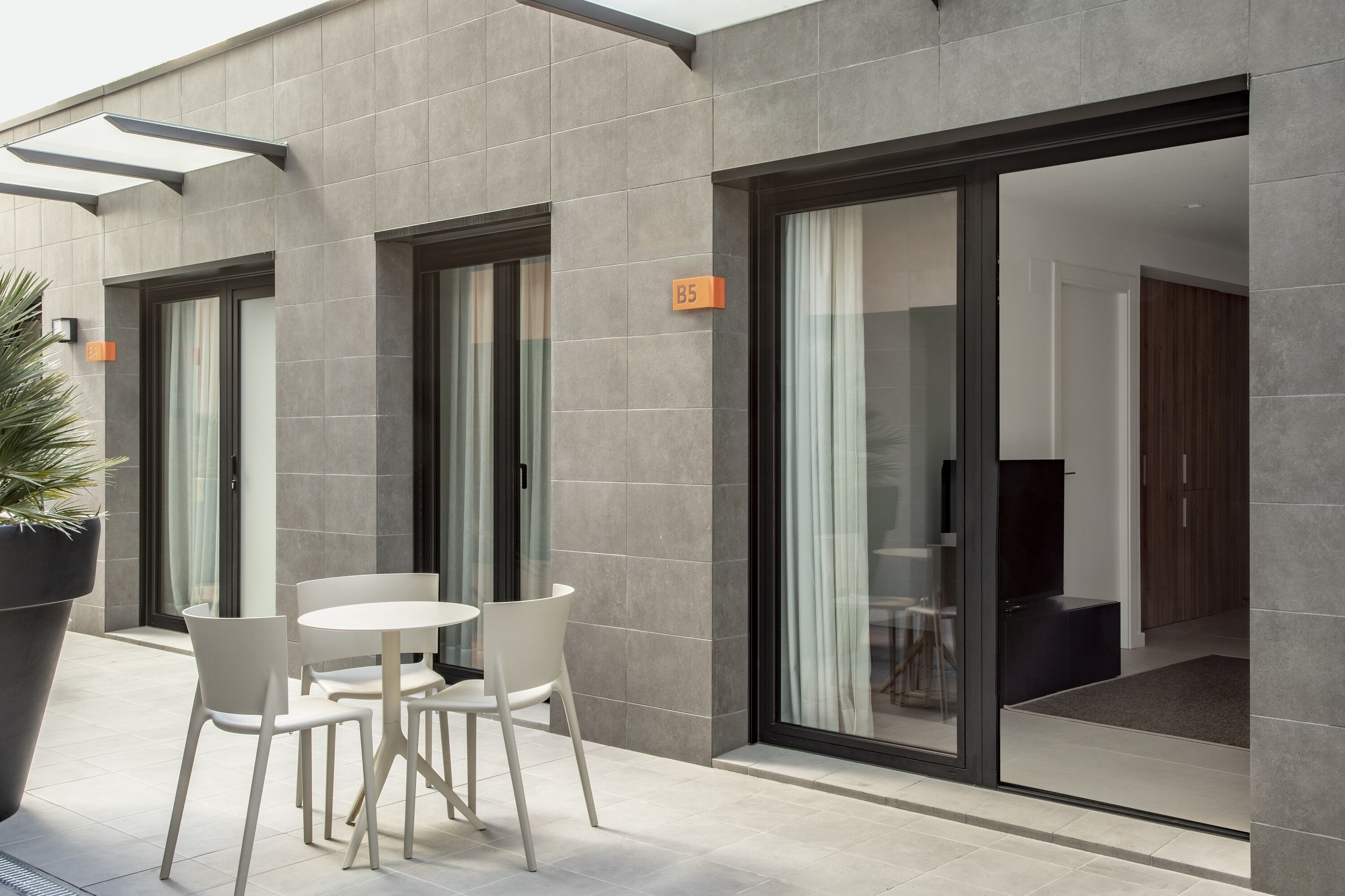 Een modern gebouw met een grijze betegelde gevel, een glazen deur met de aanduiding 'B5', en een klein buitengebied met witte stoelen en een tafel.
