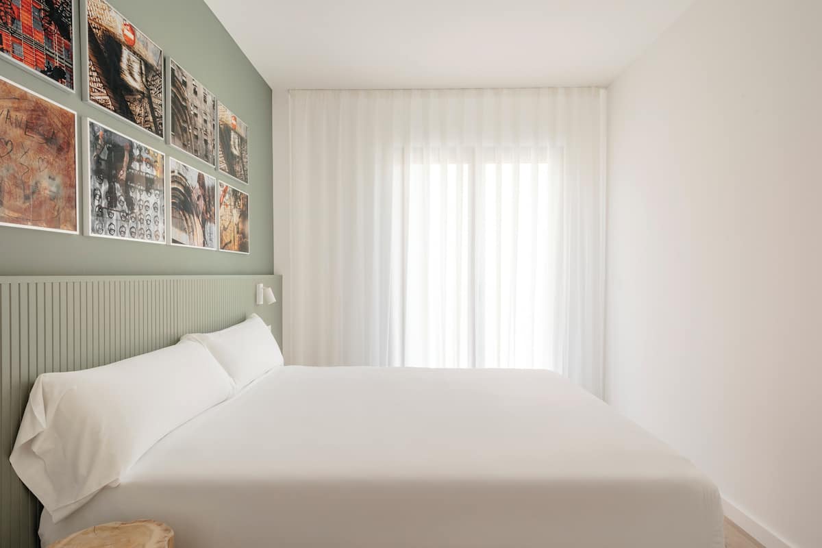 Une chambre d’hôtel avec un grand lit, une tête de lit ornée de lignes verticales, une fenêtre avec des rideaux légers et un mur décoré de plusieurs tableaux encadrés.