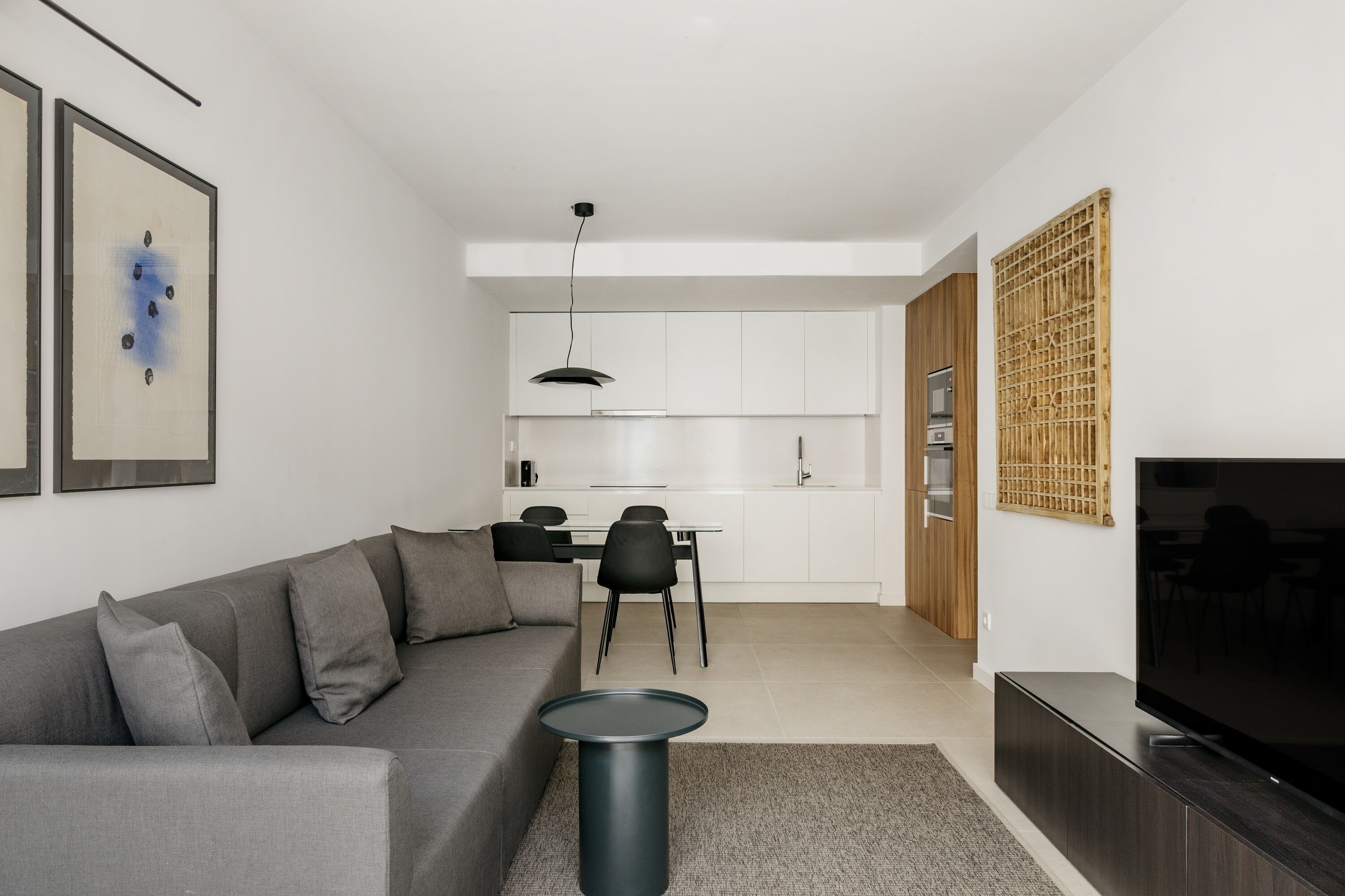 A modern living room with a grey sofa, a small round table, a flat-screen TV, and a kitchen in the background.